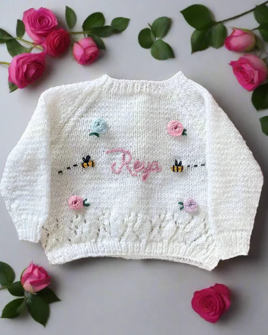White hand-knitted children's cardigan with embroidered floral and bee details from Little Flower Knits. Design a unique cardigan on our Knit Builder