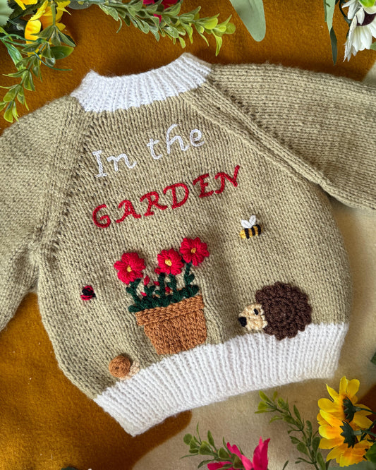 Handmade baby cardigan UK with garden theme, featuring flowers, a hedgehog, and embroidered text