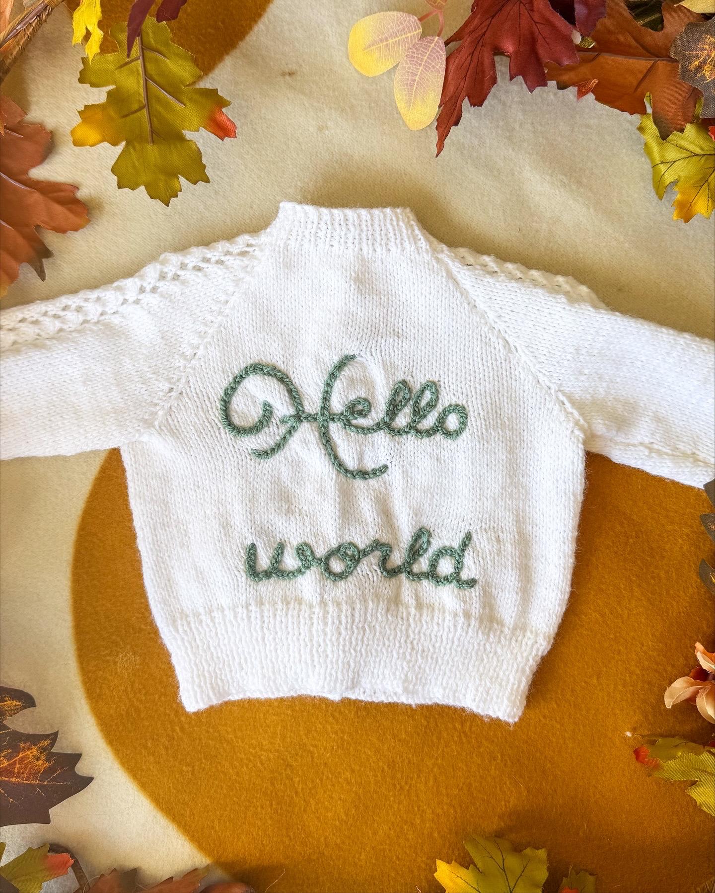 Handknitted white baby cardigan with green 'Hello world' embroidery by Little Flower Knits