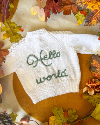 Hand-knitted baby sweater in white with 'Hello world' in green on the front by Little Flower Knits