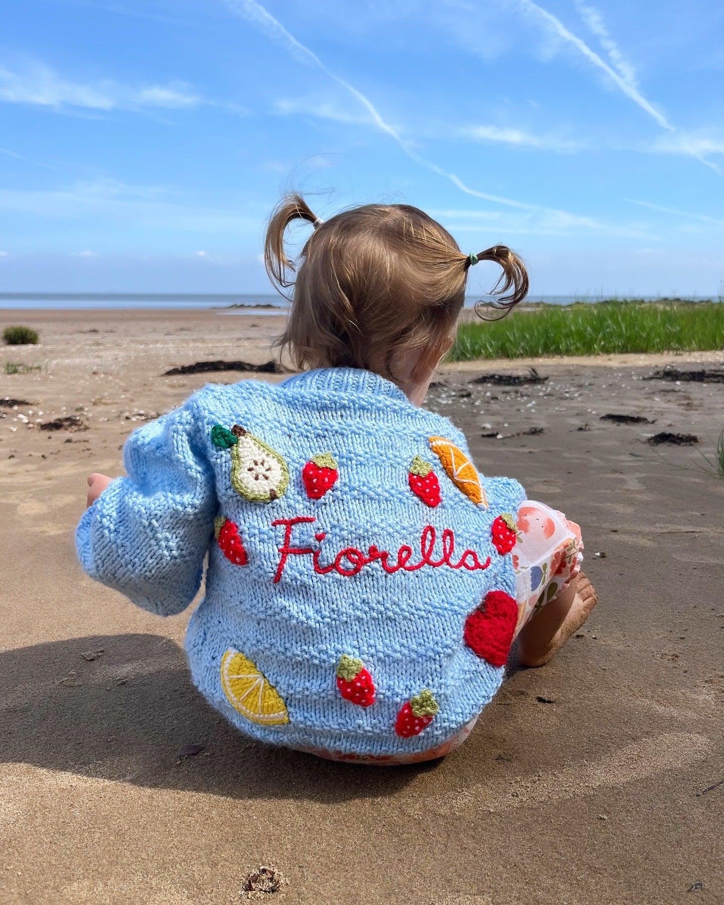 Toddler on beach in light blue fruit pattern cardigan for toddlers with colourful fruit appliques and personalised name