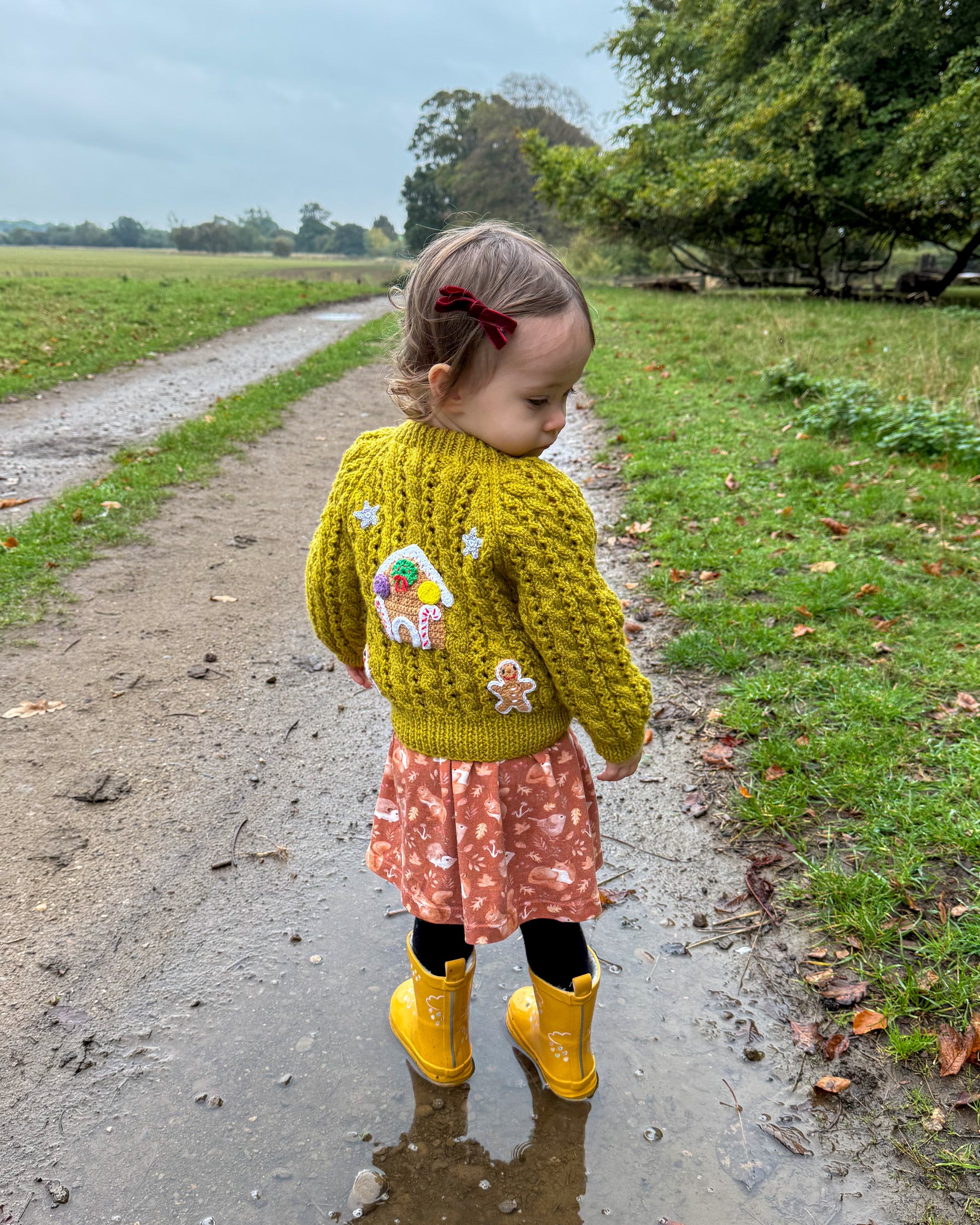 Girl wearing a gingerbread cottage baby cardigan in yellow with festive embroidery in a countryside setting