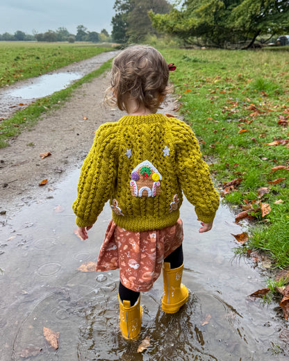 Adorable toddler wearing a gingerbread cottage baby cardigan, playing in puddles with yellow rain boots