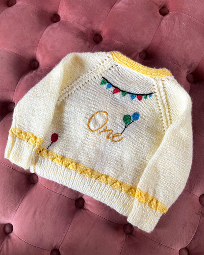 Hand knitted white and yellow baby cardigan with balloons and bunting design, unique first birthday cardigan gift