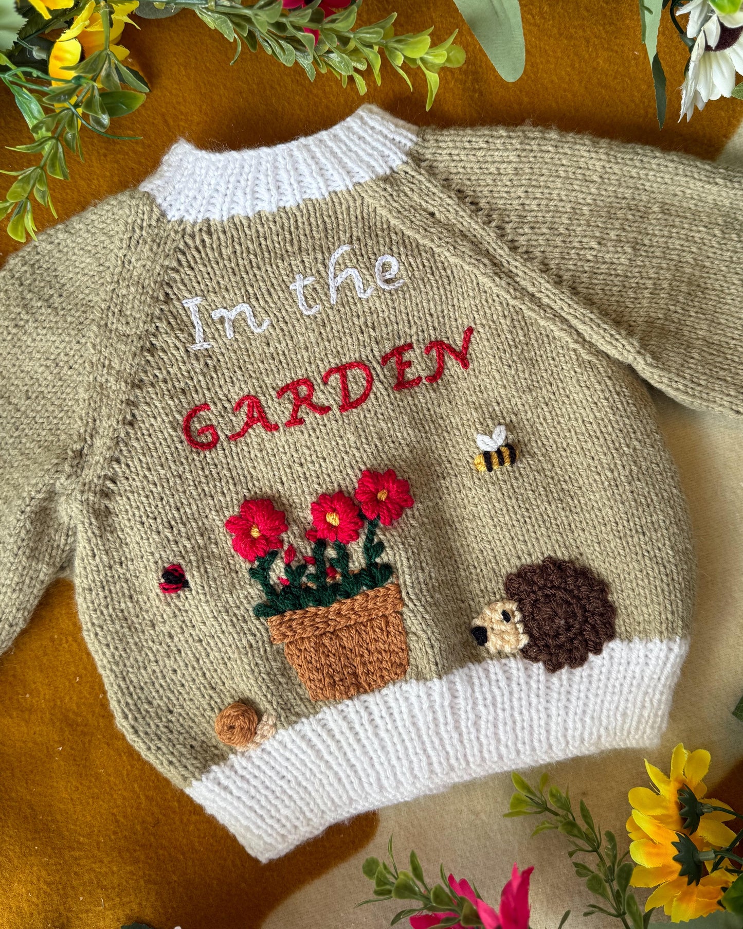 Handmade baby cardigan UK with garden theme, featuring flowers, a hedgehog, and embroidered text
