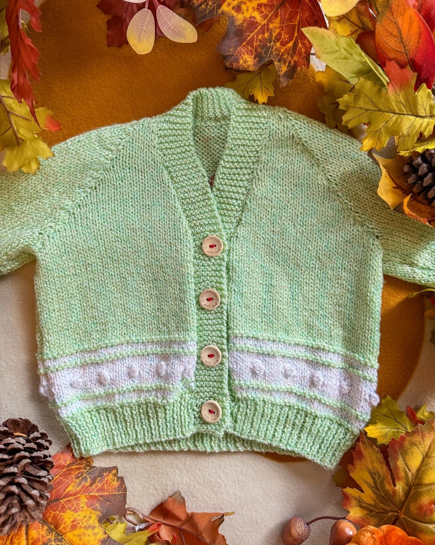 Handmade baby cardigan UK in soft green with a white trim and wooden buttons, surrounded by autumn leaves