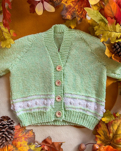 Handmade baby cardigan UK in soft green with a white trim and wooden buttons, surrounded by autumn leaves