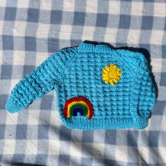 Blue newborn cardigan with rainbow and sun applique knitted by Little Flower Knits on checkered background