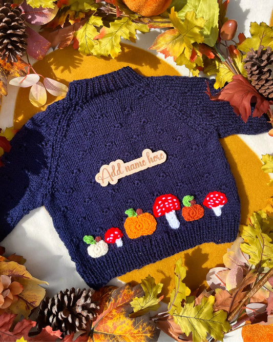 Little Flower Knits navy baby sweater with hand-knitted pumpkin and toadstool costume motif on hem and wooden name tag