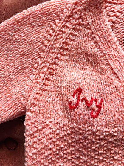 Close-up of a pink knitted personalised baby cardigan UK with the name Ivy embroidered in red