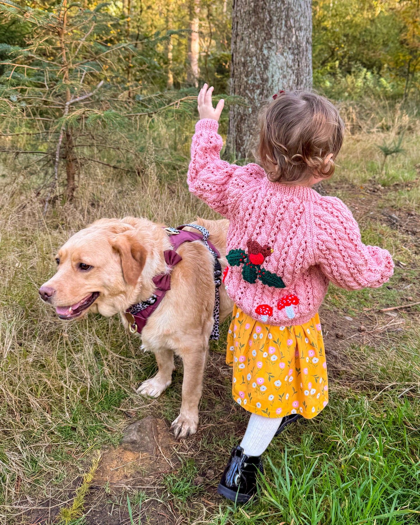 Girl wearing a pink knitted woodland themed kids cardigan with floral design, standing beside a friendly dog