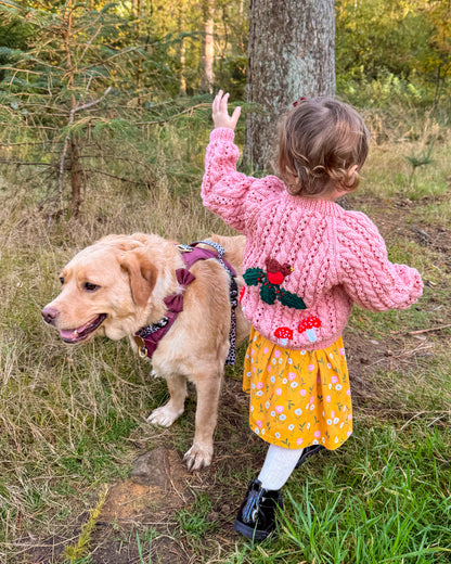 Girl wearing a pink knitted woodland themed kids cardigan with floral design, standing beside a friendly dog